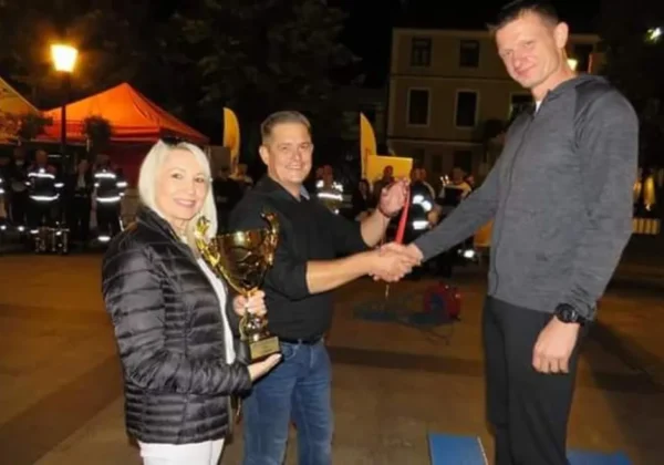 Trzy osoby na placu podczas Tonghest Firefighter Challenge Płock 2021: kobieta z pucharem, mężczyzna wręczający nagrodę i wysoki mężczyzna stojący na podium, w tle strażacy i namioty.