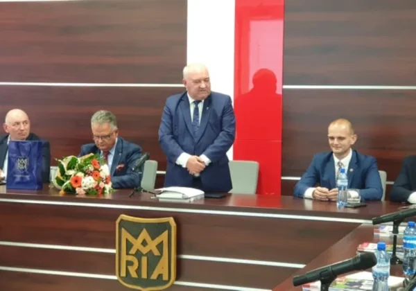 Gość na sesji Rady Gminy Bodzanów, mężczyzna w garniturze stoi przy stole, podczas gdy pozostali radni siedzą, na blacie są kwiaty, mikrofony i butelki wody, a w tle widać drewniane panele i symbol gminy.