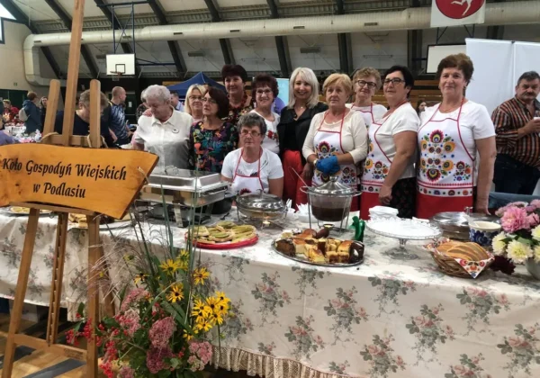 Grupa kobiet w kolorowych fartuchach z ludowymi motywami stoi za stołem z potrawami i deserami podczas festynu Pożegnanie Lata w Łącku.