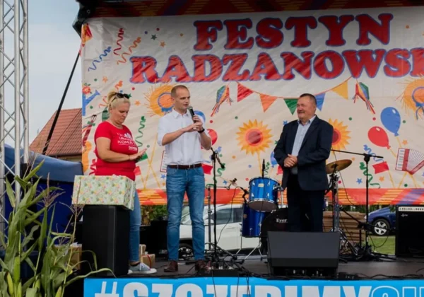 Trzy osoby stoją na scenie podczas Festynu Rodzinnego „Będzie lepiej” – mężczyzna w jasnej koszuli przemawia do mikrofonu, obok niego kobieta w czerwonej koszulce trzyma prezent, a drugi mężczyzna w garniturze uśmiecha się przy perkusji na tle kolorowego baneru.