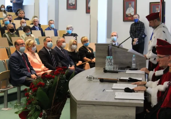 Uroczysta inauguracja roku akademickiego w Mazowieckiej Uczelni Publicznej w Płocku: uczestnicy w maseczkach siedzą w audytorium, a panel profesorów w togach zasiada przy pulpicie obok kwiatowej dekoracji.