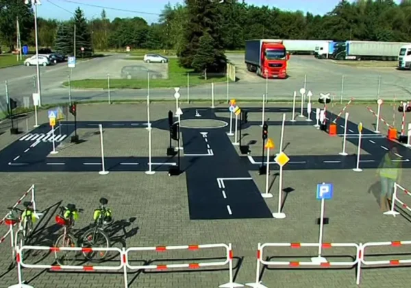 Mobilne rowerowe miasteczko drogowe na placu, z wyznaczonymi ulicami, znakami, barierkami i rowerami, służące do nauki bezpiecznego poruszania się dzieci.