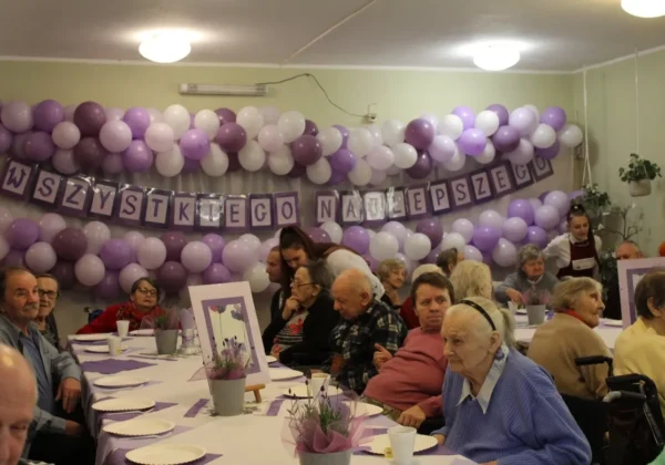 Imieniny mieszkańców Domu Pomocy Społecznej w Koszelewie – sala ozdobiona fioletowo-białymi balonami i banerem „Wszystkiego najlepszego”, przy długim stole siedzą seniorzy.