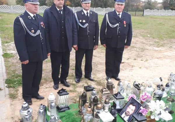 Czterech strażaków w mundurach stoi na łące obok ustawionych lampionów i fotografii w ramce, upamiętniając zmarłego druh Mariusza Bieńka.