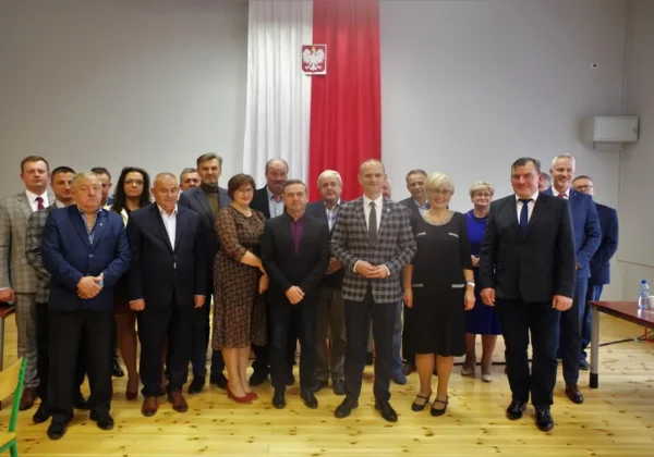 Grupa osób w garniturach i eleganckich sukienkach stoi razem na sali konferencyjnej, przed polską flagą i godłem podczas Sesji Gminy Radzanowo.