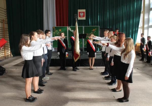 Uczniowie Zespołu Szkół im. Stanisława Staszica w Gąbinie stoją w dwóch rzędach naprzeciw siebie, wykonując gesty w stronę centralnej flagi podczas szkolnej uroczystości.