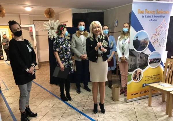 Kobieta z mikrofonem w ręku przemawia podczas proekologicznej zabawy w Domu Pomocy Społecznej Brwilno, otoczona grupą uczestników w maseczkach i dekoracjami sali.