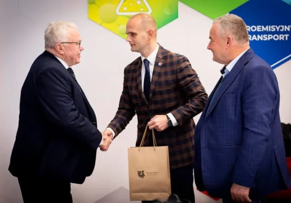 Trzech mężczyzn w garniturach wymienia uścisk dłoni podczas wizyty w PKN Orlen, a jeden z nich trzyma papierową torbę z upominkiem.