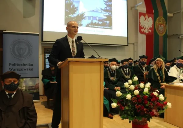Mężczyzna w garniturze przemawia z mównicy podczas inauguracji Roku Akademickiego 2021/2022 na płockiej filii Politechniki Warszawskiej, wśród zgromadzonych gości w akademickich togach i kwiatowej dekoracji.