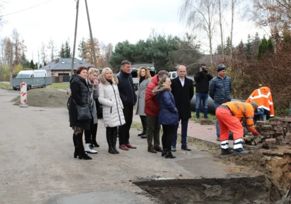 Grupa mieszkańców i urzędników stoi na poboczu drogi powiatowej, obserwując robotników w pomarańczowych odblaskowych kombinezonach pracujących przy wykopie obok stosu cegieł.