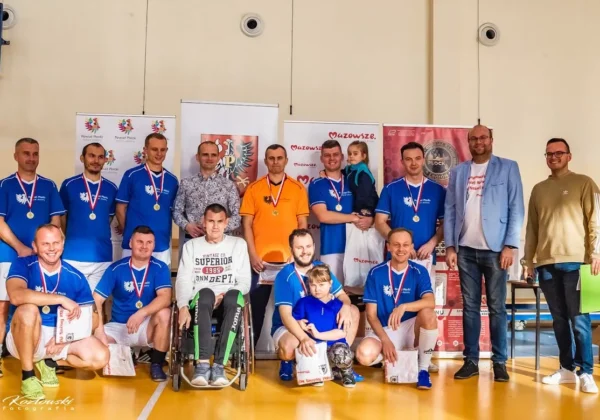 Grupa zawodników w niebieskich koszulkach z medalami na szyjach, stojących i siedzących na boisku podczas charytatywnego turnieju halowej piłki nożnej na rzecz rehabilitacji zawodnika.