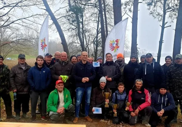 Grupa uczestników zawodów spinningowych o Puchar Starosty Płockiego stoi nad brzegiem jeziora, trzyma trofea i ma flagi w tle.