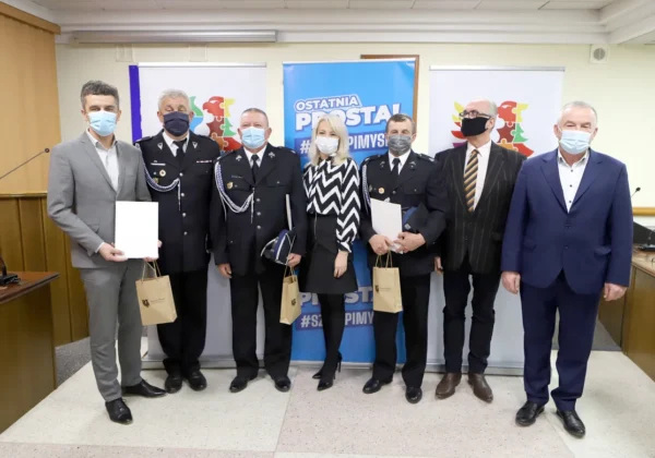 Grupa ośmiu osób w garniturach i mundurach policyjnych stoi na sali konferencyjnej przed banerami, wszyscy w maseczkach, trzymają dokumenty i torby, po podsumowaniu akcji szczepień w mobilnych punktach powiatu płockiego.