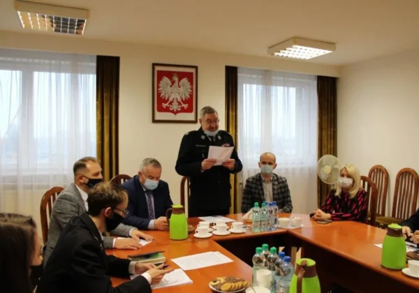 Uczestnicy posiedzenia Prezydium Zarządu Oddziału Powiatowego ZOSP RP w Płocku siedzą przy stole w sali konferencyjnej, a stojący w mundurze strażackim mężczyzna odczytuje dokument; na ścianie widnieje godło Polski.
