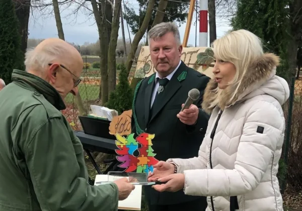 Trzy osoby podczas obchodów 60-lecia Koła Łowieckiego Kampinos — kobieta w białej kurtce wręcza mężczyźnie w zielonej marynarce upominek, podczas gdy drugi mężczyzna trzyma mikrofon i prowadzi przemówienie.