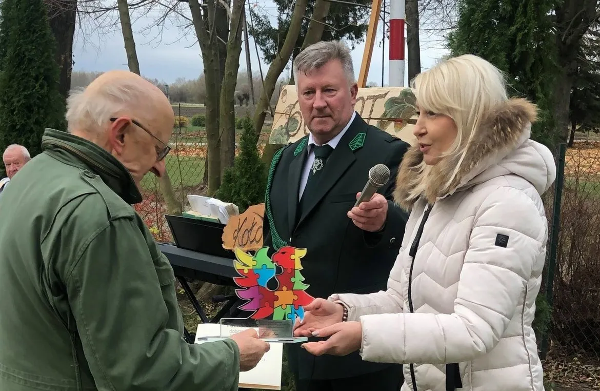 Trzy osoby podczas obchodów 60-lecia Koła Łowieckiego Kampinos — kobieta w białej kurtce wręcza mężczyźnie w zielonej marynarce upominek, podczas gdy drugi mężczyzna trzyma mikrofon i prowadzi przemówienie.