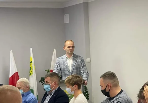 Starosta Płocki przemawia z mównicy podczas sesji Rady Gminy Staroźreby; obok siedzą radni, niektórzy w maseczkach, w tle flagi.