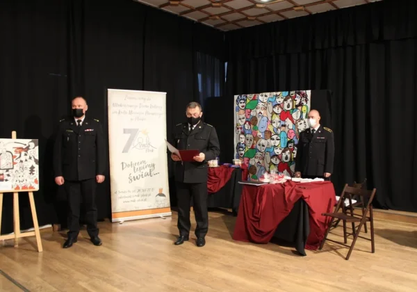 Trzy osoby w mundurach stoją na scenie podczas powiatowo-miejskich eliminacji konkursu „Czujka na straży Twojego bezpieczeństwa”, na tle widoczne są baner i kolorowy mural z twarzami.