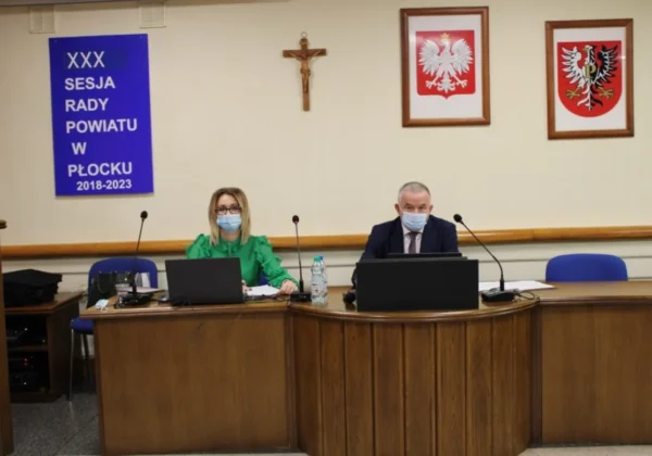 Dwóch urzędników Rady Powiatu w Płocku siedzi przy wspólnym biurku z laptopami i mikrofonami podczas XXX sesji, ponownie w trybie zdalnym, a na ścianie wiszą krzyż i dwa herby.