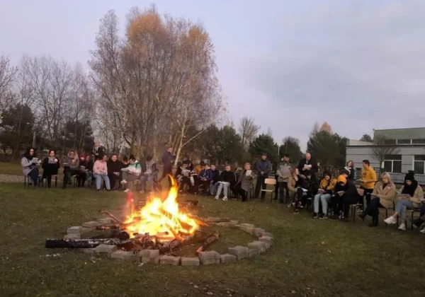 Grupa osób siedzi na zewnątrz wokół ogniska otoczonego kamiennymi bloczkami podczas wydarzenia „Światełko dla Niepodległej” w Zespole Szkół im. Leokadii Bergerowej w Płocku.