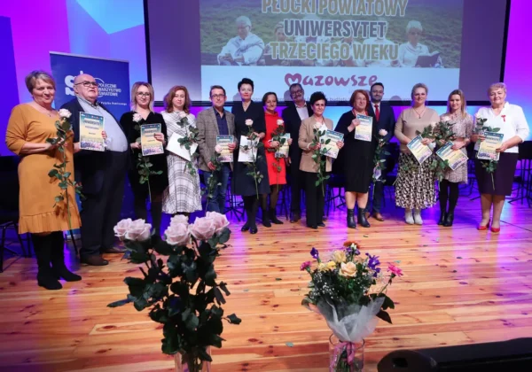 Grupa osób stoi na scenie podczas jubileuszowej inauguracji roku akademickiego Płockiego Powiatowego Uniwersytetu Trzeciego Wieku, trzymając dyplomy i róże, a w tle widać ekran z napisem wydarzenia.