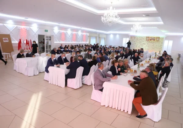 Duża grupa osób siedzi przy długich stołach nakrytych białymi obrusami podczas uroczystości Niepodległości w jasnej sali z żyrandolami i flagami w tle.
