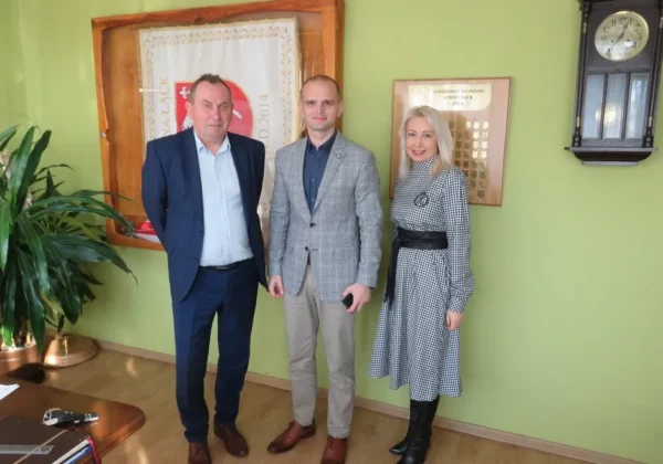 Trójka urzędników Powiatu Płockiego podczas roboczej wizyty w Gminie Łąck stoi w zielonym pomieszczeniu biurowym, obok roślin i tablic pamiątkowych.