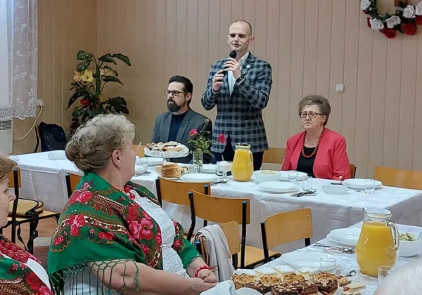 Mężczyzna przemawia do zgromadzonych seniorów podczas spotkania w Łęgu Probostwie, stojąc przy stole z napojami i przekąskami.