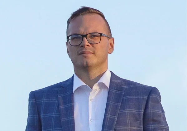 Portret nowego wójta gminy Radzanowo, mężczyzny w granatowym garniturze w kratę i białej koszuli, stojącego na tle błękitnego nieba.