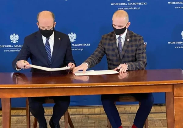 Dwóch mężczyzn w garniturach i maseczkach siedzi przy drewnianym stole, podpisując dokumenty dotyczące modernizacji jedenastu przejść dla pieszych w Powiecie Płockim, na niebieskim tle z napisem Wojewoda Mazowiecki.