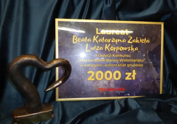 Statuetka w kształcie serca stoi obok tablicy informacyjnej z laureatami XI edycji konkursu Mazowieckie Barwy Wolontariatu i nagrodą 2000 zł w kategorii wolontariat grupowy.