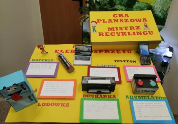 Żółty stół edukacyjny z grą planszową „Mistrz recyklingu” oraz rekwizytami elektronicznymi (laptop, drukarka, telefon, bateria, akumulator) ilustrujący naukę poprzez odkrywanie i dociekanie.
