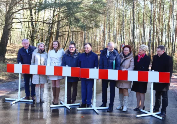 Grupa osób w zimowych ubraniach stoi za czerwono-białą barierą na mokrej drodze podczas uroczystego otwarcia wyremontowanej drogi powiatowej Soczewka – Łąck w lesie.