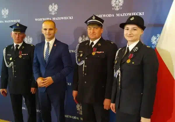 Na zdjęciu znajduje się czterech mężczyzn i kobiet, ubranych w mundury strażackie, którzy pozują do zdjęcia z odznaczeniami Brązowego Krzyża Zasługi w uroczystej atmosferze.