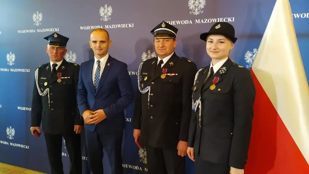 Na zdjęciu znajduje się czterech mężczyzn i kobiet, ubranych w mundury strażackie, którzy pozują do zdjęcia z odznaczeniami Brązowego Krzyża Zasługi w uroczystej atmosferze.