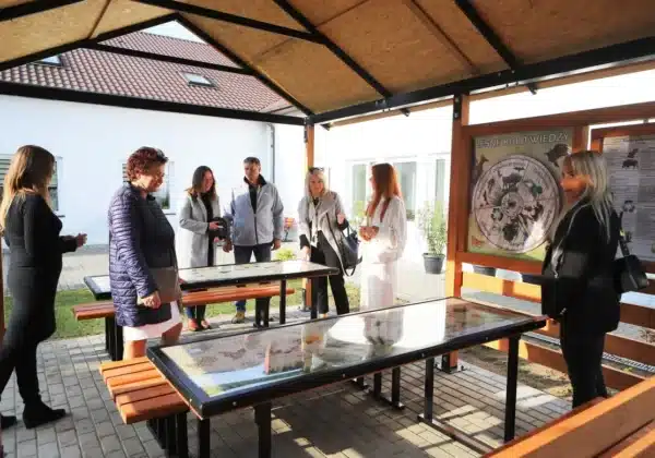 Na zdjęciu przedstawiono nowe wiaty ekologiczne w Domu Pomocy Społecznej w Koszelewie, gdzie grupka osób dyskutuje przy stolikach poświęconych tematyce przyrodniczej.