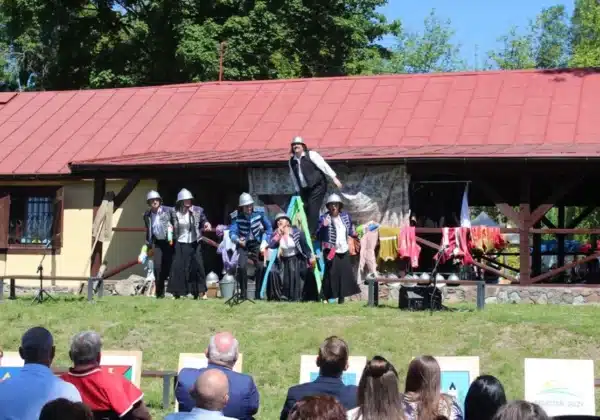 Na zdjęciu przedstawiono występ artystyczny z okazji 100-lecia Ochotniczych Straży Pożarnych w powiecie płockim, z aktorami w mundurach i kostiumach na tle wiejskiego budynku.