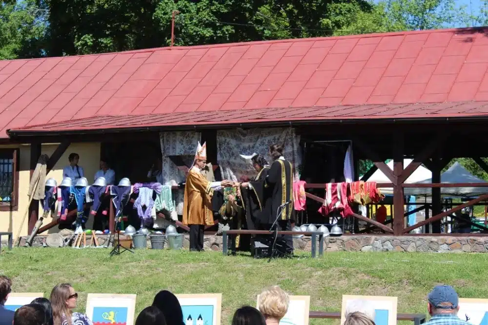 Uroczystość z okazji 100-lecia Ochotniczych Straży Pożarnych w powiecie płockim, na której uczestnicy stoją przed budynkiem, ubrani w tradycyjne stroje.