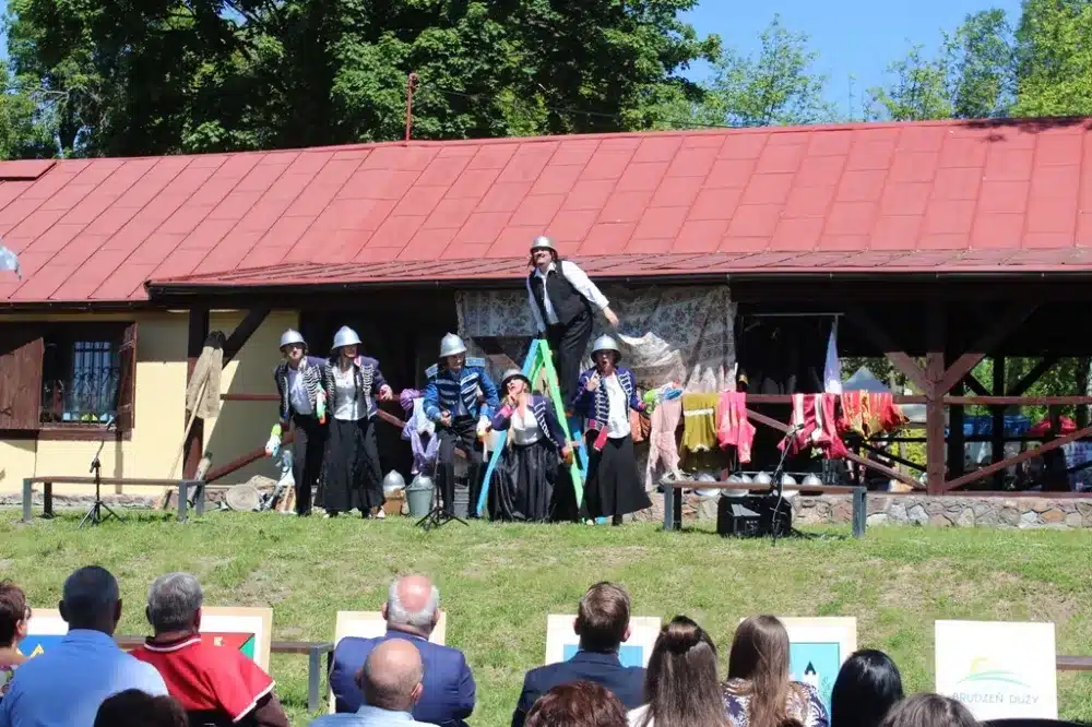 Na zdjęciu przedstawiono występ artystyczny z okazji 100-lecia Ochotniczych Straży Pożarnych w powiecie płockim, z artystami w strojach ludowych, wykonującymi akrobatyczne układy na tle malowniczej scenerii.