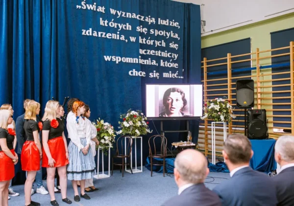 Grupa uczniów i gości stoi na scenie podczas jubileuszu 100-lecia Zespołu Szkół im. Leokadii Bergerowej w Płocku, przed ekranem z czarno-białym zdjęciem i kwiatami, na tle niebieskiej kurtyny z napisem.