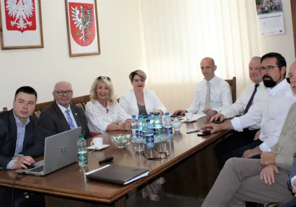 Grupa urzędników i współpracowników siedzi przy długim stole konferencyjnym podczas posiedzenia Zarządu Powiatu Płockiego, z laptopem, dokumentami i butelkami wody na stole, a na ścianach wiszą herby państwowe.