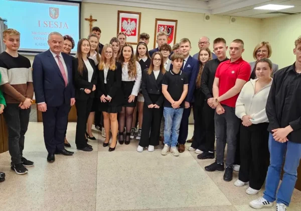 Grupa młodych radnych i urzędników stoi razem w sali sesyjnej podczas I sesji Młodzieżowej Rady Powiatu w Płocku (kadencja 2024–2026), z ekranem projekcyjnym z napisem „I SESJA” i godłami państwa w tle.