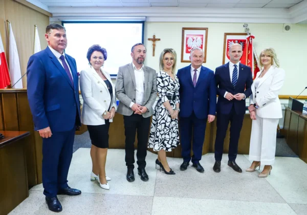 Siedmioosobowa grupa radnych i urzędników w formalnych strojach stoi w sali sesyjnej Powiatu podczas I sesji kadencji 2024–2029, z flagami państwowymi i herbami w tle.