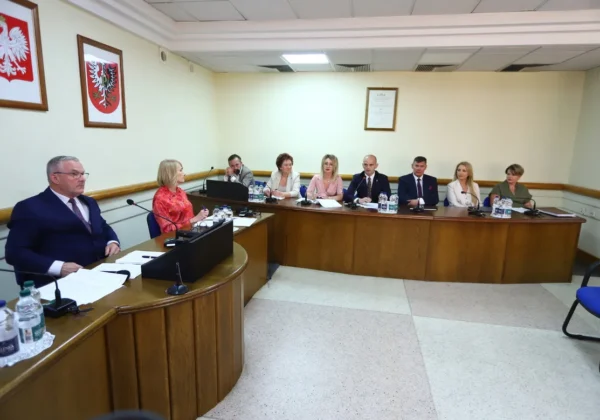 Grupa radnych siedzi przy półokrągłym stole w sali sesyjnej podczas II sesji Rady Powiatu w Płocku, z mikrofonami i dokumentami na stole, a na ścianach widoczne godła państwowe.
