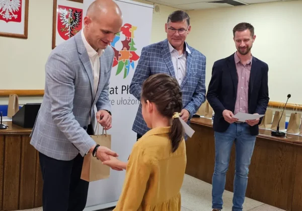 Podczas ceremonii wręczania nagród w konkursach Ligii Ochrony Przyrody mężczyzna w jasnoszarej marynarce przekazuje upominek dziewczynce w żółtym ubraniu, gdy pozostali dwaj mężczyźni obserwują; w tle widoczne są banery i godła.