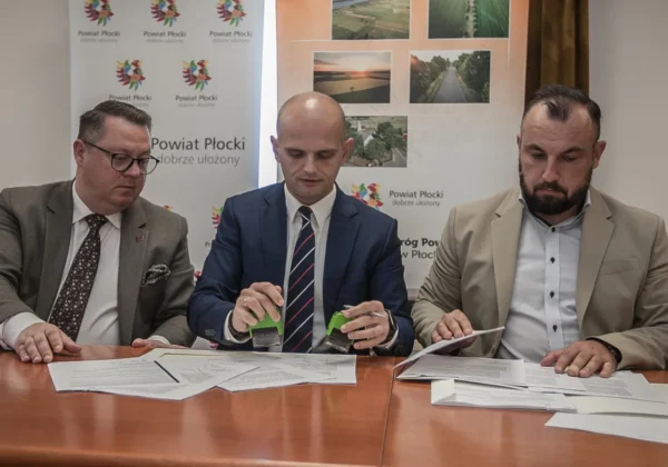 Trzech mężczyzn w garniturach siedzi przy stole i podpisuje umowy na przebudowę dróg powiatowych, na stole leżą dokumenty, a w tle widoczne są banery Powiatu Płockiego.