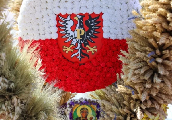 Konkursowy wieniec dożynkowy z białych i czerwonych kwiatów otoczony kłosami zbóż, z centralnym godłem Polski na czerwonej tarczy oraz ikoną Matki Bożej.