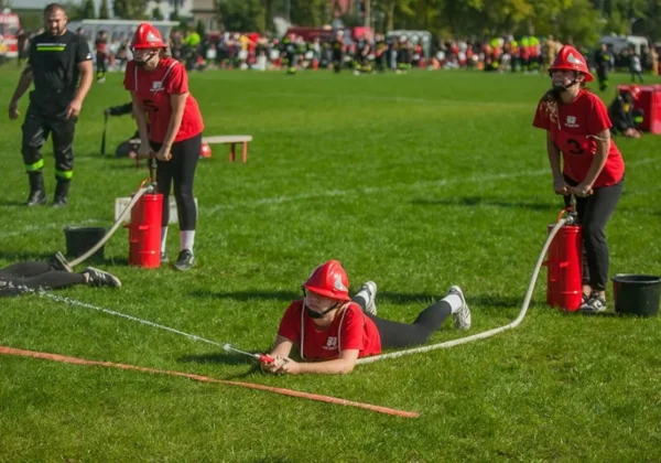 Trzy osoby w czerwonych koszulkach i hełmach strażackich ćwiczą z wężem gaśniczym na zielonej trawie podczas XIII Powiatowych Zawodów Sportowo-Pożarniczych OSP i MDP.