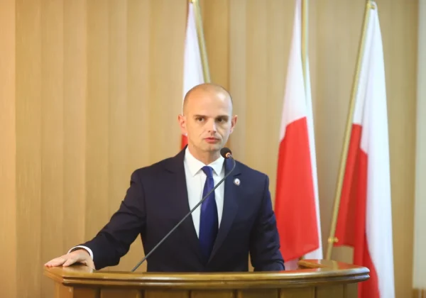 Mężczyzna w garniturze przemawia z mikrofonem zza mównicy podczas IV sesji Rady Powiatu w Płocku, w tle widoczne flagi.