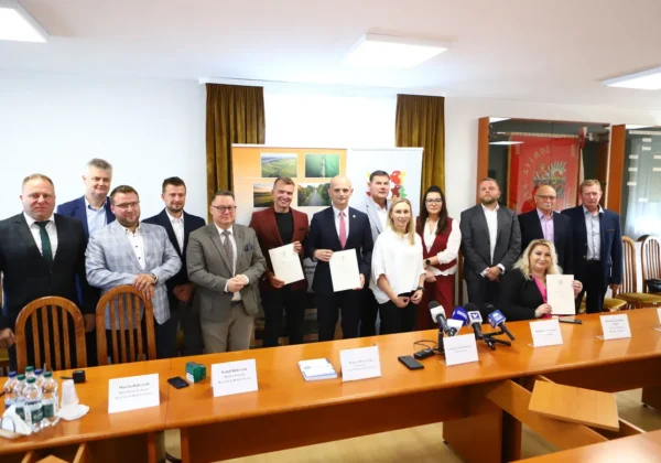 Grupa osób w garniturach stoi przy długim stole podczas konferencji prasowej, prezentując dokumenty dotyczące nowej drogi i dwóch mostów.